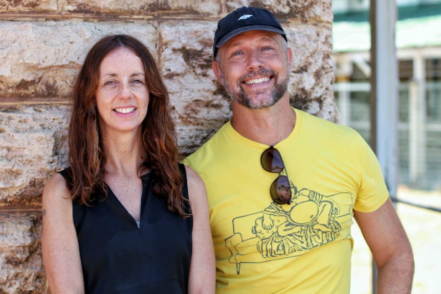 Maxine Doyle and Paul Selwyn-Norton leaning against a brick wall and smiling