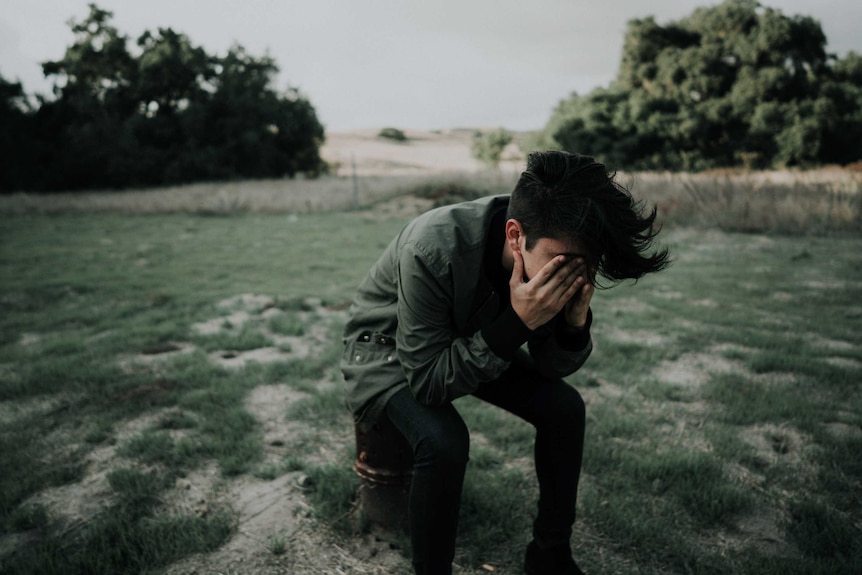 A person sits outdoors with their head in their hands, behind them is a large yard, trees and hills.