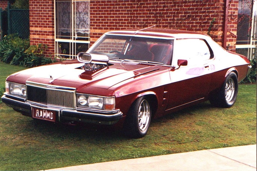 A maroon Holden Monaro classic car parked on grass in front of a red brick house