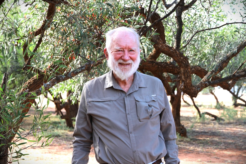 94-year-old man with white beard and hair standing in front of trees and on red dirt -- wearing a long sleeve grey shirt
