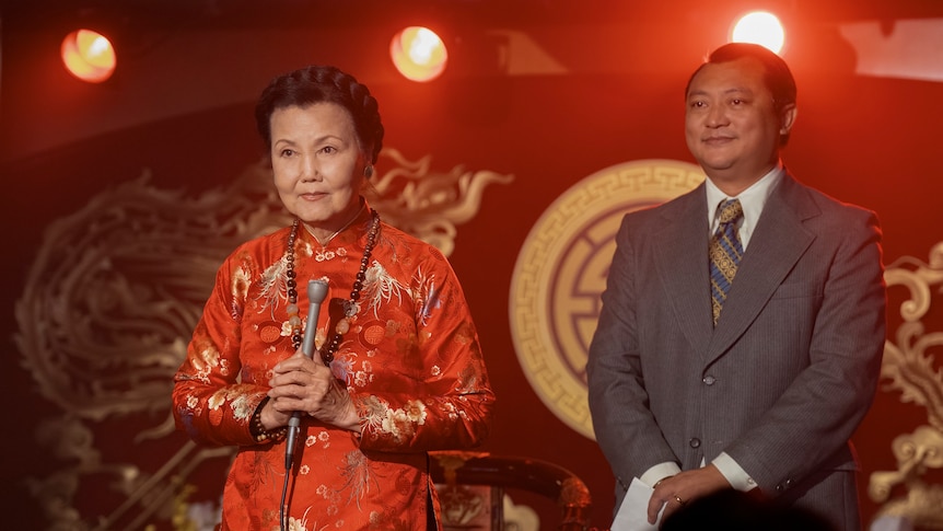 A film still showing an older woman with black hair and a middle-aged man, both Vietnamese, on a stage with a red background