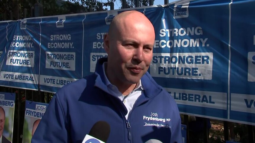 Josh Frydenberg wears blue jacket bearing his name in front of campaign posters during last federal election.