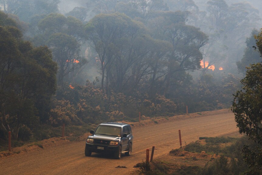 Car on dirt road with bushfire visible in scrub nearby.