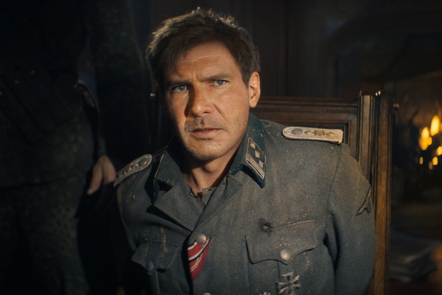 A middle-aged white man with ashen hair wears a dust-covered military uniform and is tied to a chair near a fireplace.