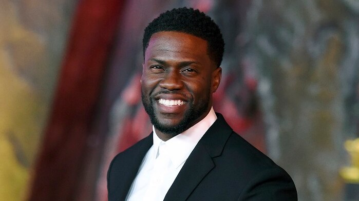 Kevin Hart, wearing a suit, smiles and holds up two fingers as he poses for the camera while arriving at a movie premiere.