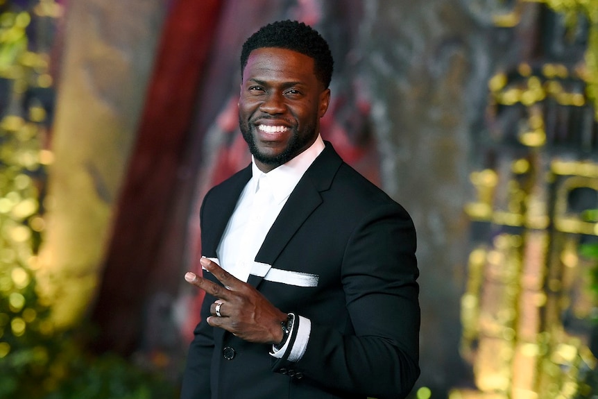 Kevin Hart, wearing a suit, smiles and holds up two fingers as he poses for the camera while arriving at a movie premiere.
