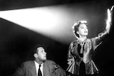 An elegant woman stands in the light of a projector as a seated man looks on.