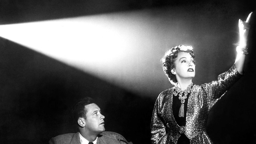 An elegant woman stands in the light of a projector as a seated man looks on.