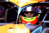 F1 driver Oscar Piastri lowers himself into his orange McLaren F1 car, wearing his full race suit, including a helmet.
