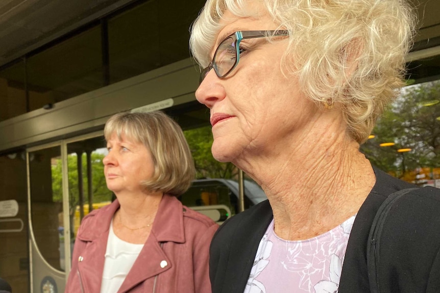 Two older women speak to microphones on a city street