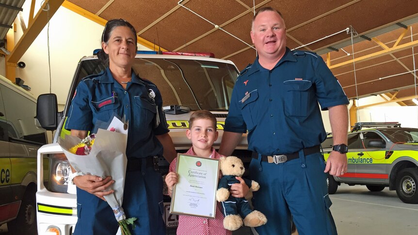 Two paramedics stand with a young boy who is happy and holding a bravery certificate.