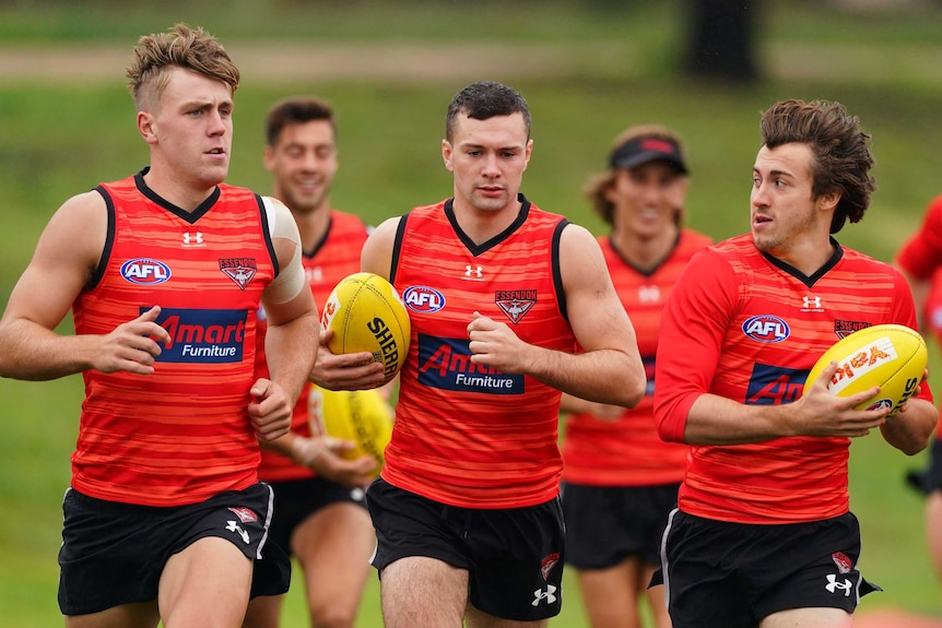 Conor McKenna holds a yellow AFL ball under his right arm, wearing a red singlet with teammates all around him
