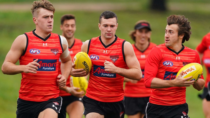 Conor McKenna holds a yellow AFL ball under his right arm, wearing a red singlet with teammates all around him