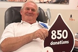 older man with white hair sitting in chair donating blood holding sign 