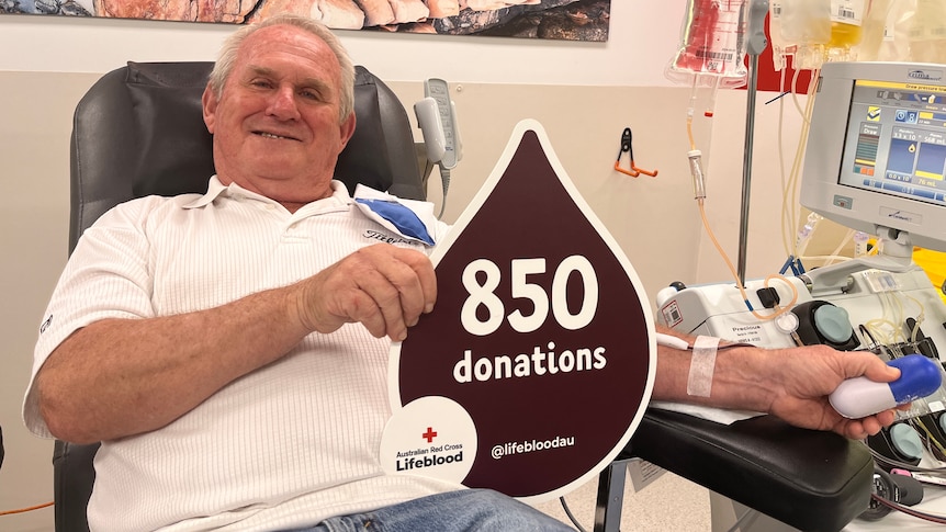 older man with white hair sitting in chair donating blood holding sign 