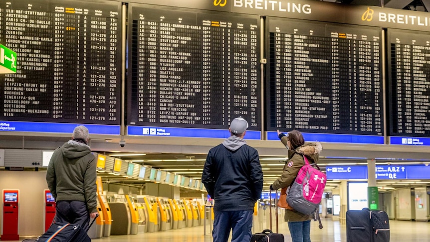 An board displaying international flights is looked at by three passengers holding luggage.