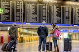 An board displaying international flights is looked at by three passengers holding luggage.