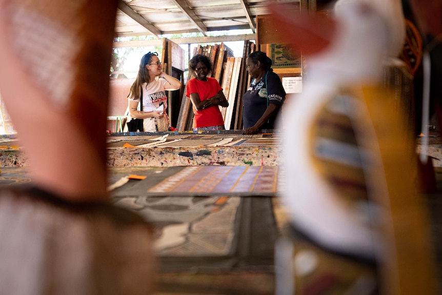 A person talks to two artists at an art centre, there are carvings in the forefront and paintings in from of them.