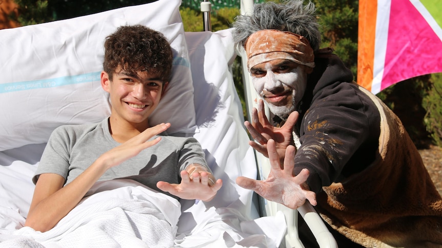 A young boy in a hospital bed holds his hands towards the camera next to an aboriginal man with face paint. 