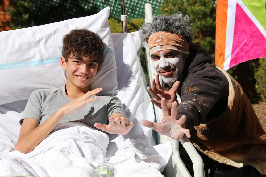 A young boy in a hospital bed holds his hands towards the camera next to an aboriginal man with face paint. 