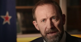 Andrew Little looks to the camera mid-speech. He wears a black suit, white shirt and patterned tie. A NZ flag is in background.