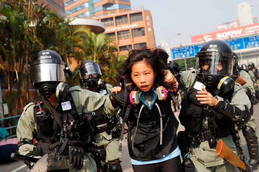 Two riot police with helmets, masks and breathing equipment pull a female protester down the street.
