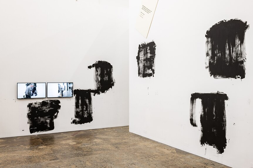 Two side by side tv screens showing Mike Parr performance hang near various roughly painted black squares on white gallery wall.