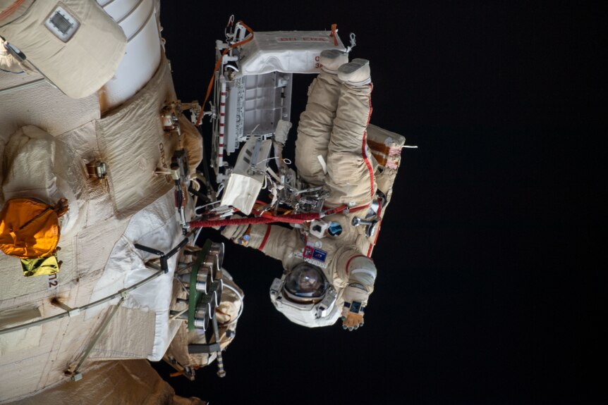Une personne portant une combinaison d'astronaute salue la caméra, photographiée à l'envers dans un espace noir