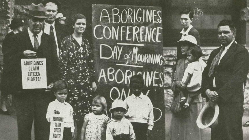 Indigenous protesters gather around a placard  that reads "Aborigines conference - day of mourning"