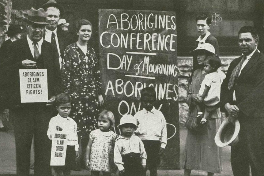 Indigenous protesters gather around a placard  that reads "Aborigines conference - day of mourning"