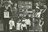 Indigenous protesters gather around a placard  that reads "Aborigines conference - day of mourning"