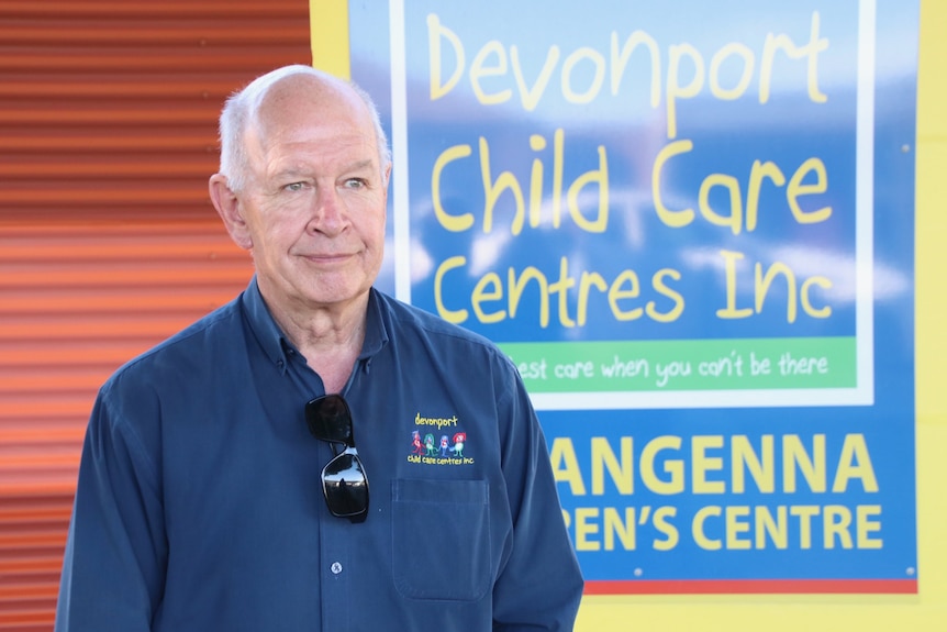 A man in a blue work shirt stands in front of a sign that reads 'Devonport Child Care Centres Inc'.