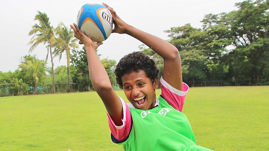 A girl in pink school uniform and green rugby bib flies high to catch a rugby ball over her head.