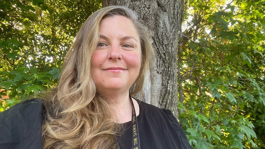 A fair-skinned Aboriginal woman with blonde hair standing in front of a tree. She is smiling with kindness.