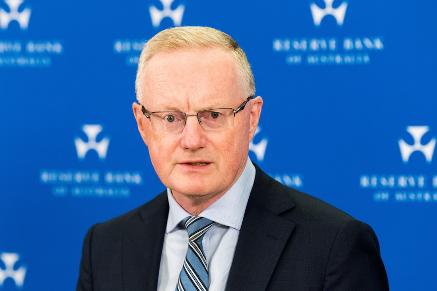 An older man with greying light hair and glasses wearing a suit and tie stands in front of a blue Reserve Bank of Australia wall