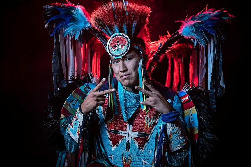 A man in vibrant feathered black, blue and white Native American regalia poses with hands in double peace sign pose.