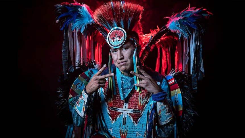 A man in vibrant feathered black, blue and white Native American regalia poses with hands in double peace sign pose.