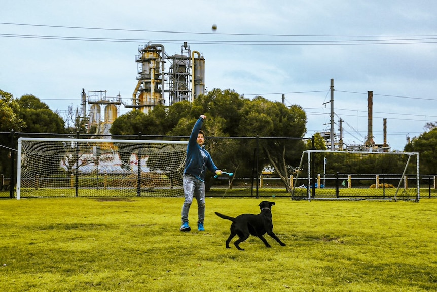 A man plays with his dog in a field in front of a refinery.