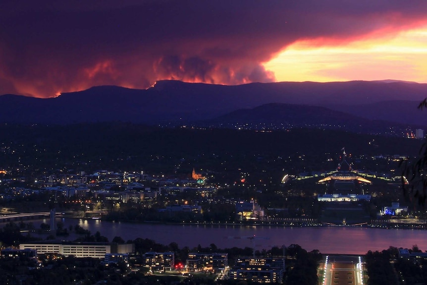 A city with a lake at sunset, and larges plumes of smoke rising from fires on the mountain ridge in the background.