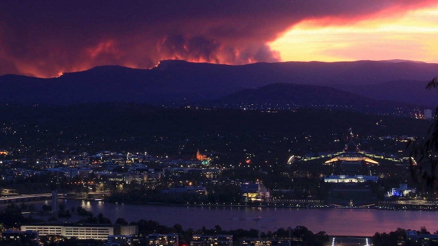 A city with a lake at sunset, and larges plumes of smoke rising from fires on the mountain ridge in the background.