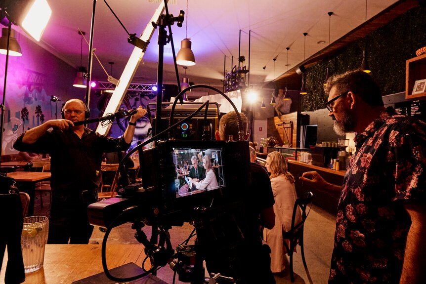 Cameras and lighting equipment surround actors inside a dimly-lit bar.