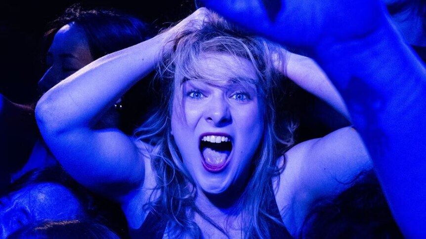 blonde haired woman hands on head with mouth open as if screaming in blue light with other human arms raised around her