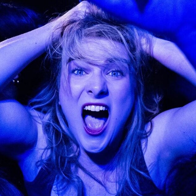 blonde haired woman hands on head with mouth open as if screaming in blue light with other human arms raised around her