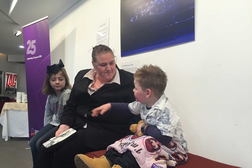 Gayle Hullah sits on a bench in the foyer of a theatre with her daughter and son