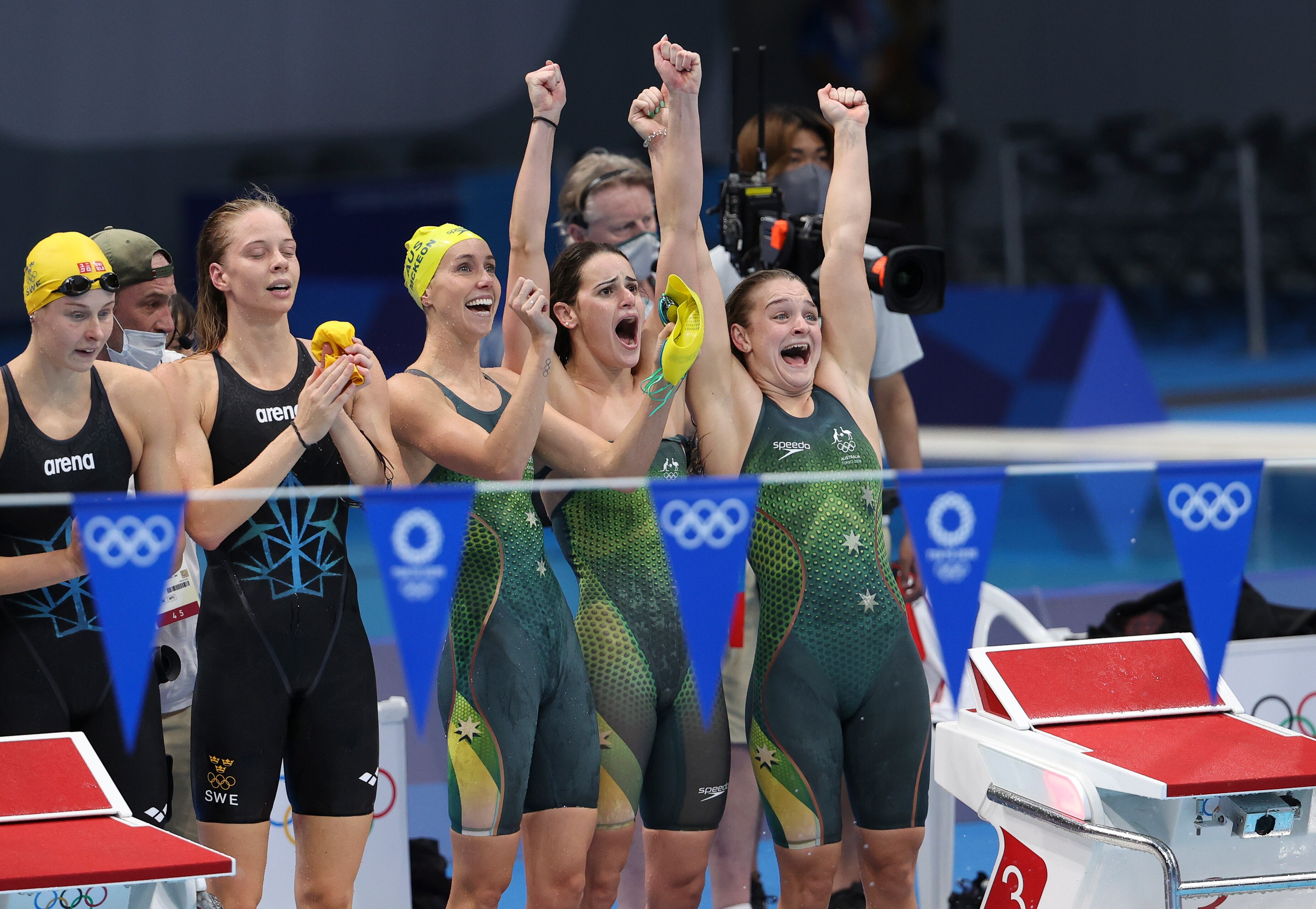 a group of swimmers cheer on their teammates from the side of the pool waving their hands in the air