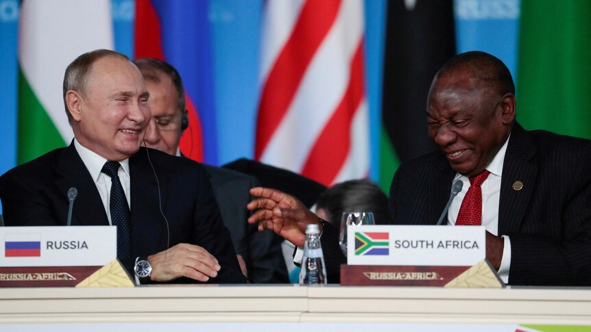 Vladimir Putin and Cyril Ramaphosa laugh together while seated in front of a row of flags.