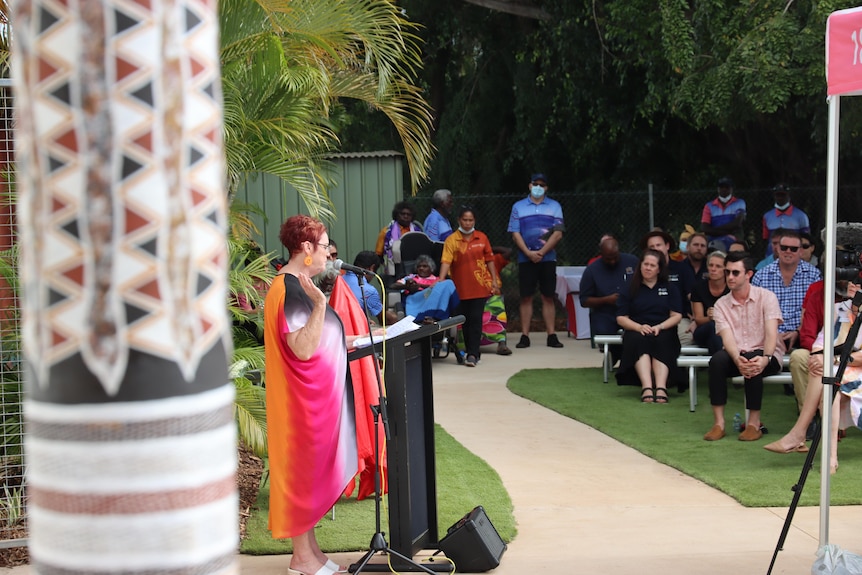 A woman standing at a lectern outside and speaking to a group of people sitting on chairs. 