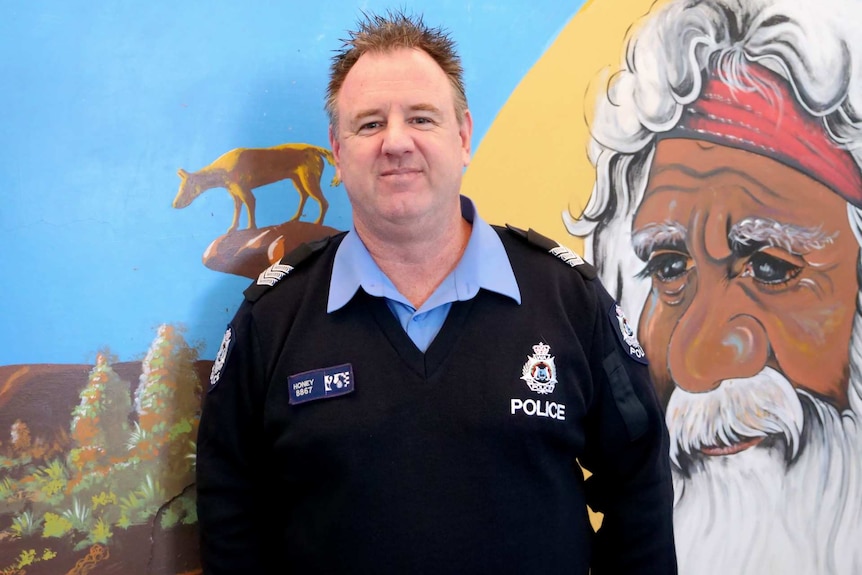 A police officer stands in front of a bright mural of an Indigenous elder looking over a landscape.