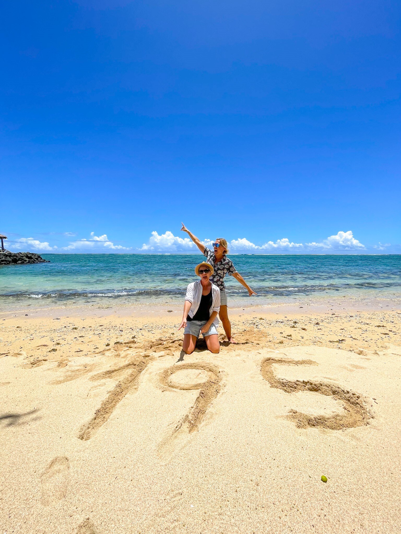 Two women on the beach jumping around a 195 sign drawn into the sand 