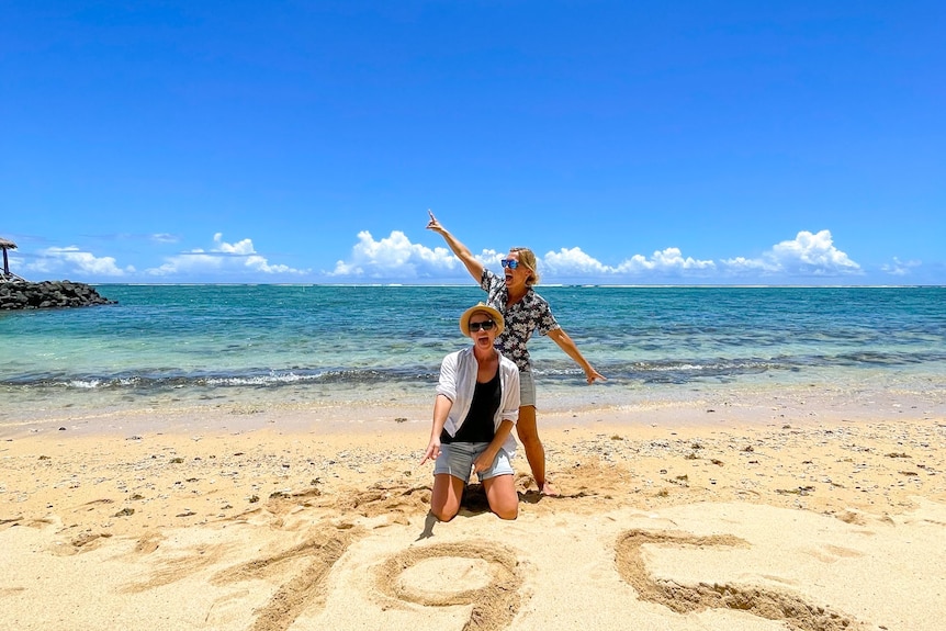 Two women on the beach jumping around a 195 sign drawn into the sand 